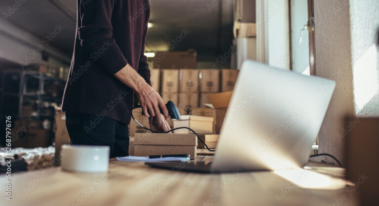 Person packing a box in a workspace with laptop and morning sunlight.